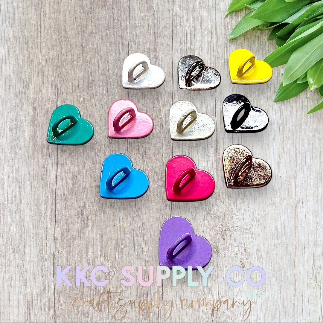 A collection of heart-shaped hooks in various colors arranged on a light wood background. The colors include green, pink, blue, purple, white, yellow, red, black, and metallic shades. A cluster of green leaves is positioned at the top right corner, adding a natural touch. The KKC Supply Co logo in pastel colors overlays the bottom of the image.